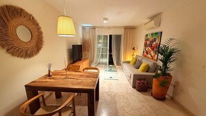 Un salon comprenant une table à manger en bois, des chaises, un téléviseur, un canapé avec des coussins, une plante en pot et un grand miroir accroché au mur.