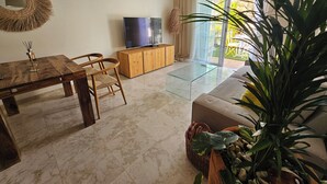 Un salon comprenant une table à manger en bois, des chaises, une table basse en verre, un téléviseur posé sur un meuble en bois et une grande plante en pot.