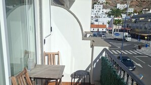 A balcony with a wooden table and chairs, a potted plant, and a view of a street with buildings and a mountain in the background. - Mogan Playa 19 by El Sirocco (Mogan)