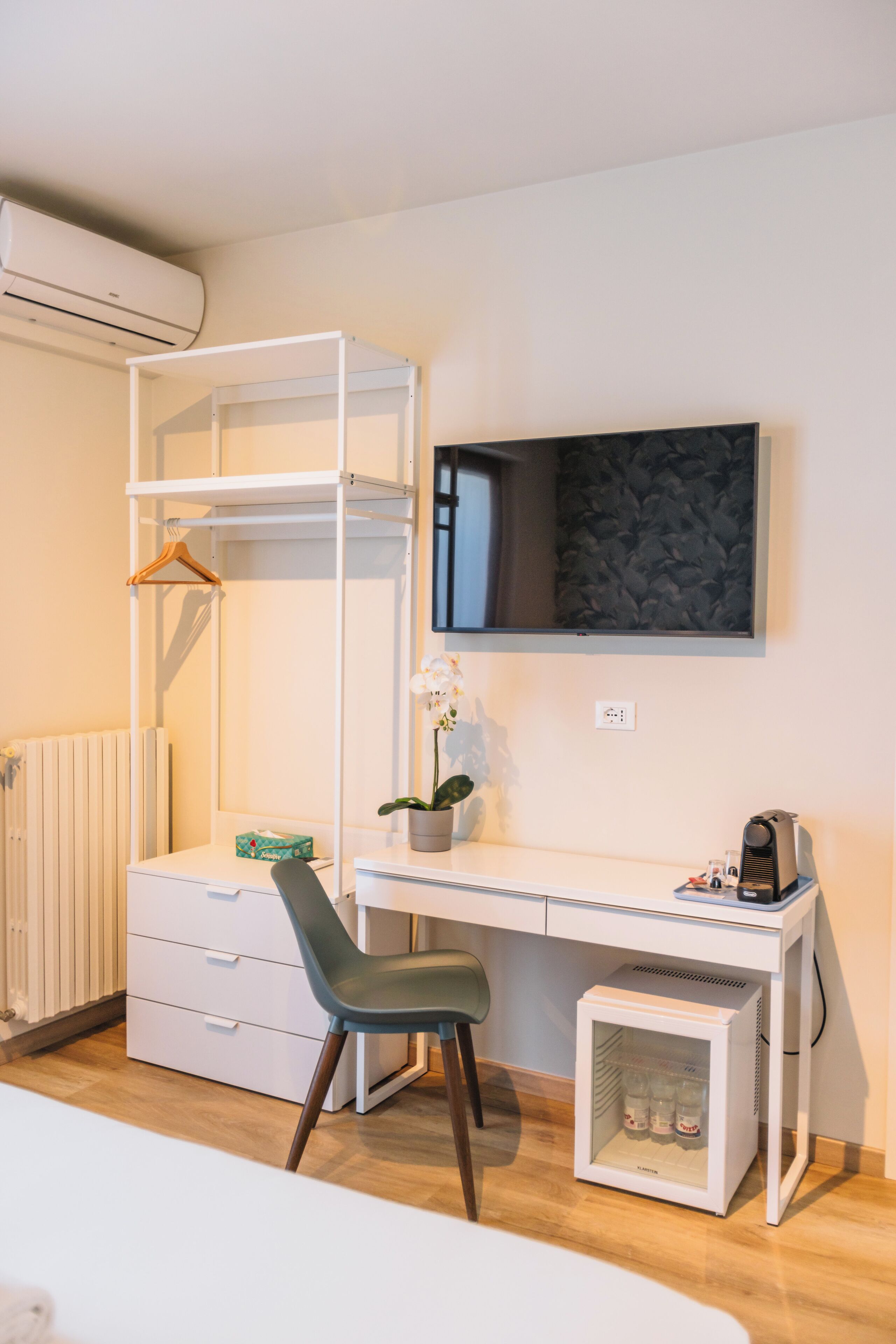 Une pièce de taille modeste, comprenant un bureau blanc, une chaise, un petit réfrigérateur, un radiateur et un téléviseur fixé au mur.