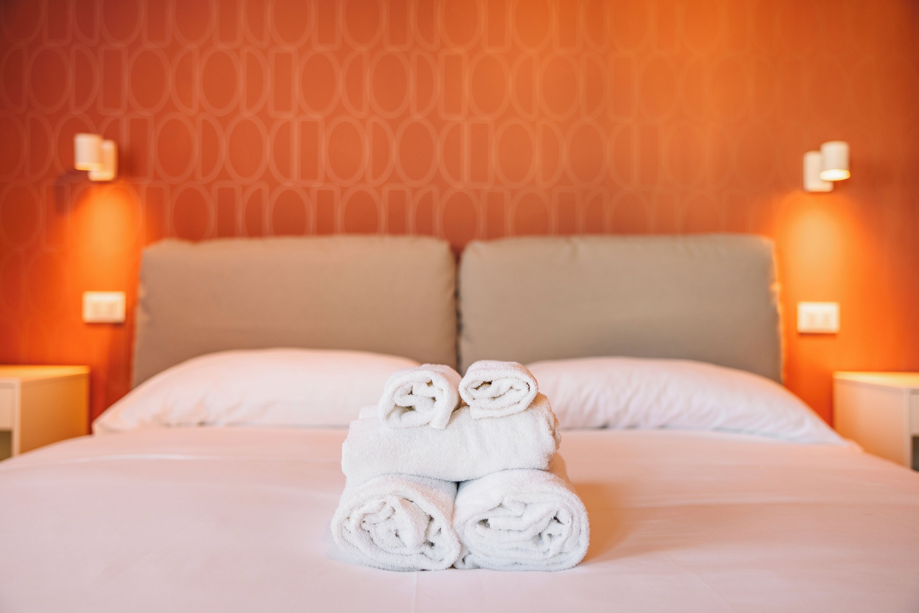 A neatly made bed with rolled white towels in the center, a patterned wall, and two bedside lamps.