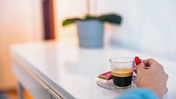 Een hand die een kop koffie vasthoudt op een witte tafel, met een wazige achtergrond inclusief een plant.