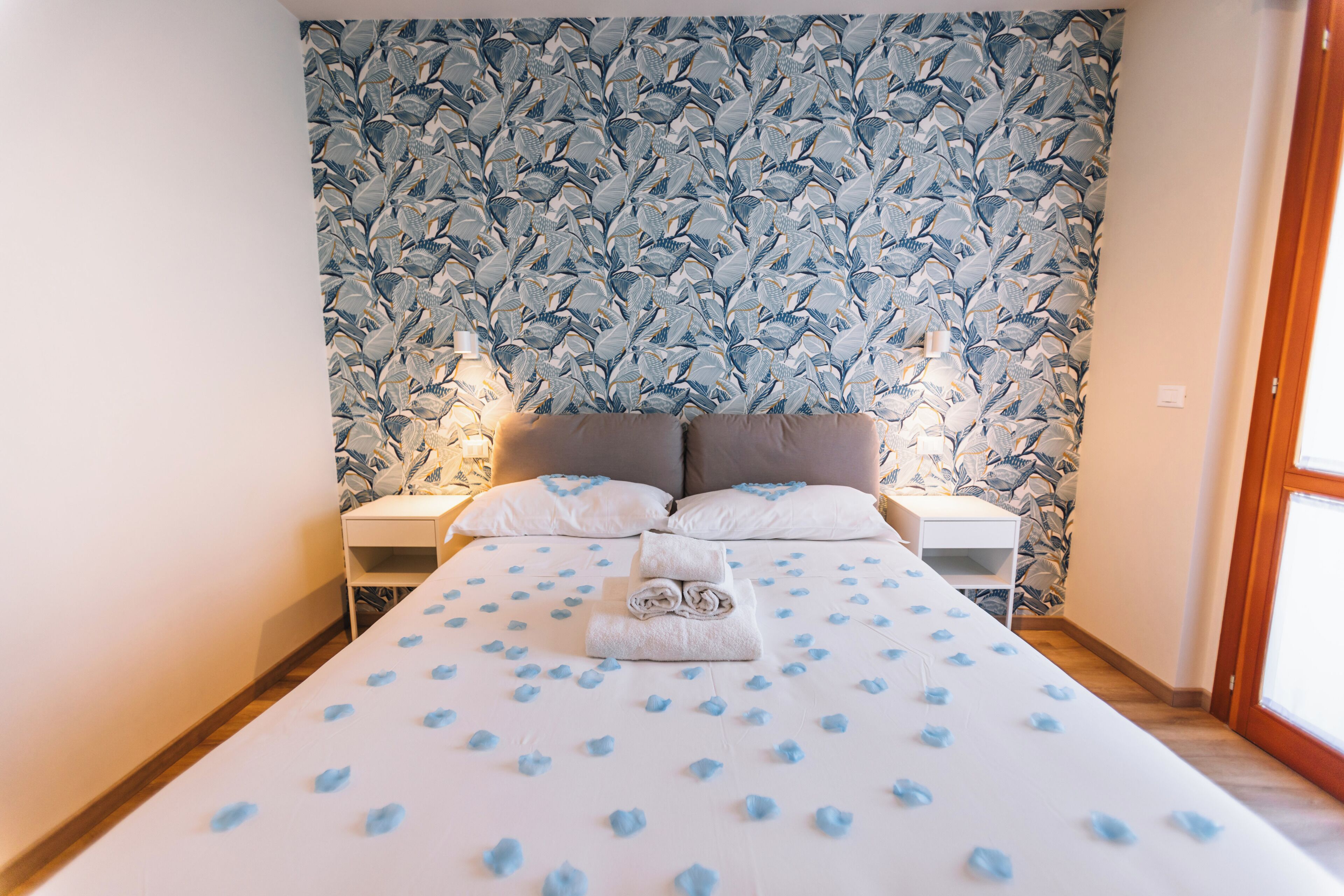 A neatly made bed with a white bedspread and blue floral wallpaper. There are two bedside tables with lamps, a white nightstand, and a window on the right.