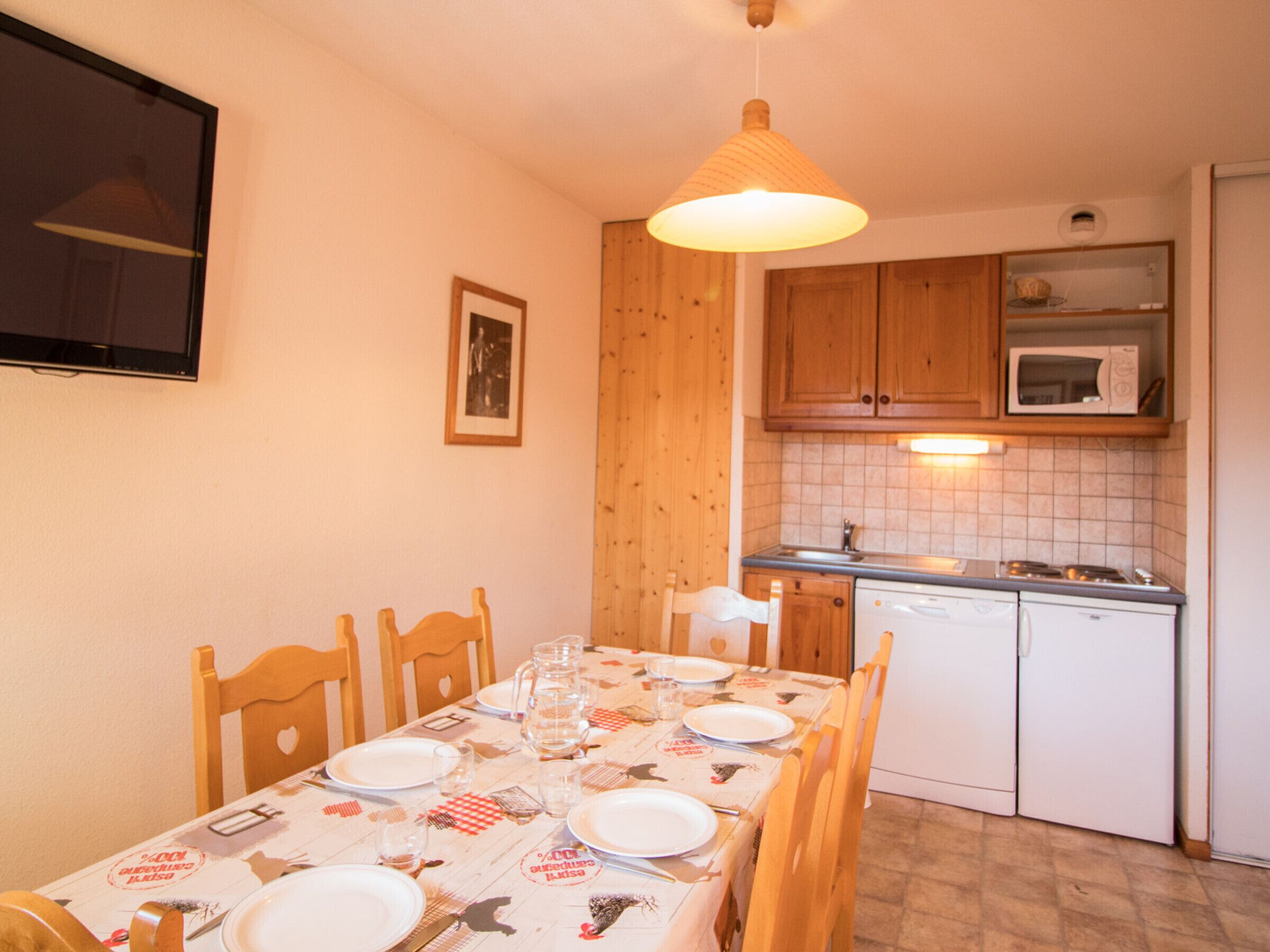 A dining area with a table set for four, wooden chairs, a hanging light fixture, and a kitchen with wooden cabinets and a microwave.