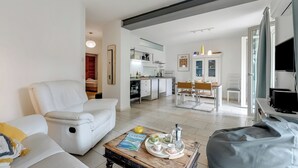 Een moderne woonkamer met een witte bank, een salontafel met een tijdschrift en een grijze loungestoel. Op de achtergrond is een keuken met witte kasten en een eethoek met een tafel en stoelen te zien.