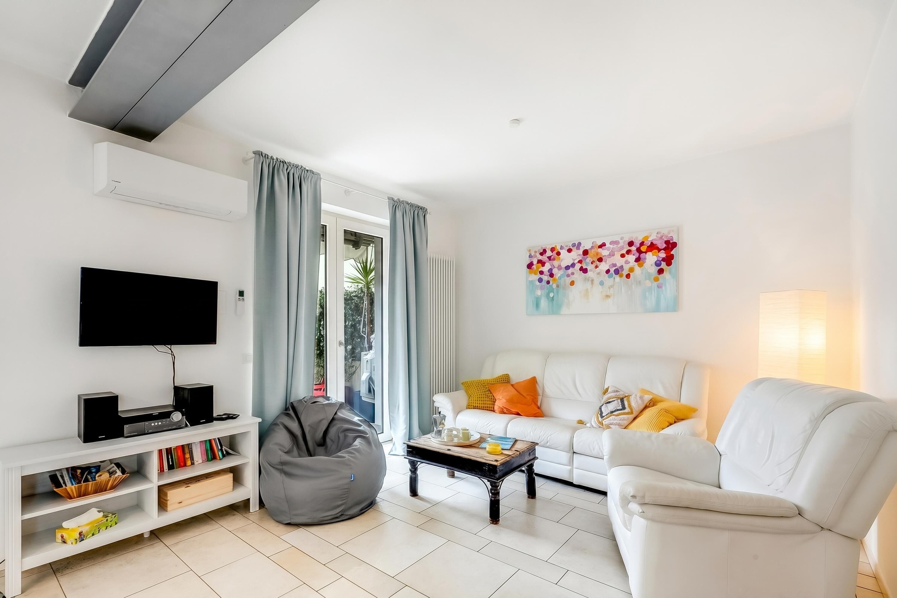 A modern living room with a flat-screen TV, a white sofa with colorful cushions, a coffee table, a bean bag, and a painting on the wall.