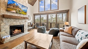 A spacious living room with a stone fireplace, large windows, and a cozy seating area. - Camp Care Free at Eagles Nest - game room, hot tub, fire pit (Banner Elk)
