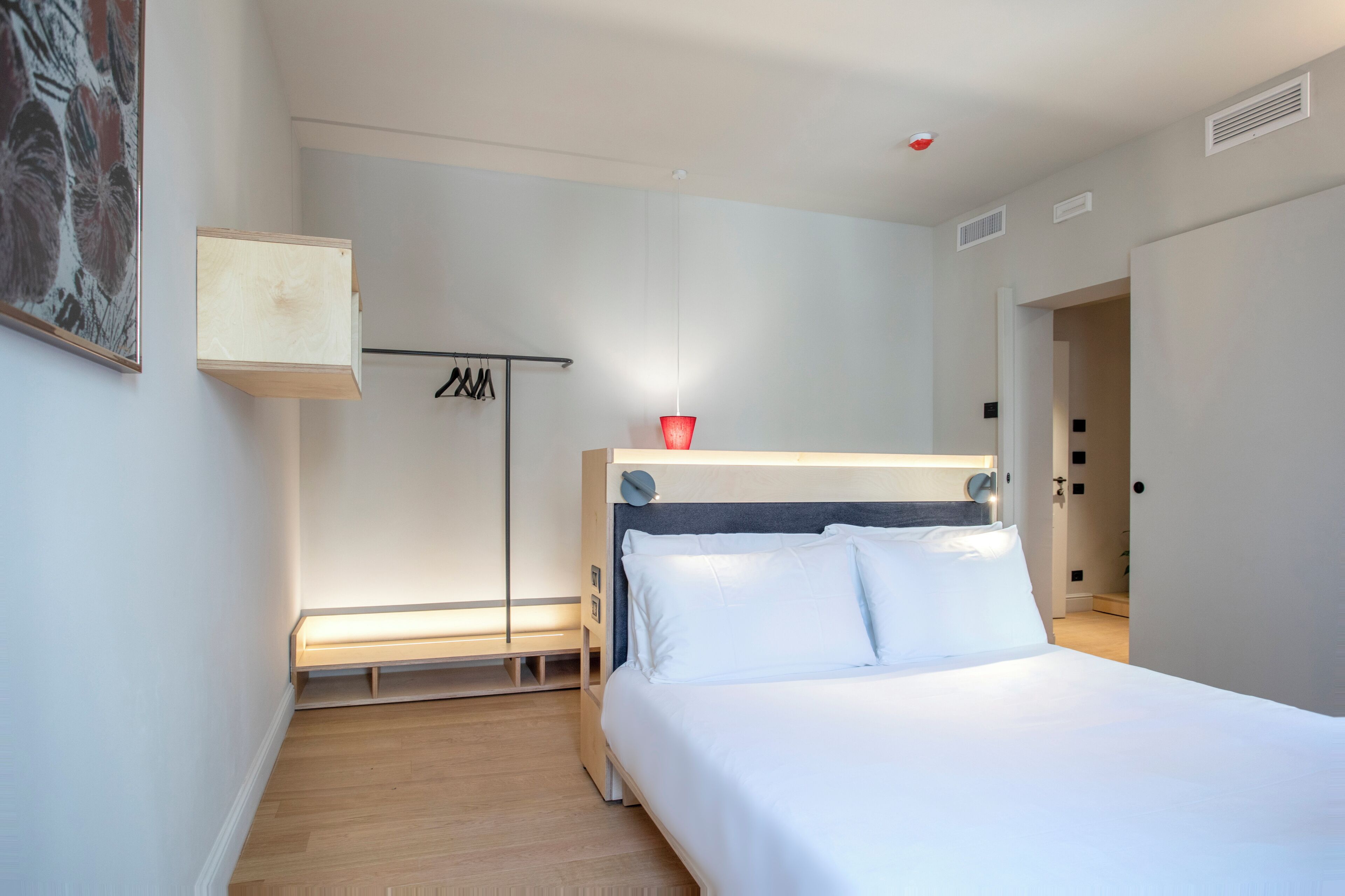 A modern bedroom with a wooden floor, a bed with white linens, a bedside table, a hanging light, and a coat rack.