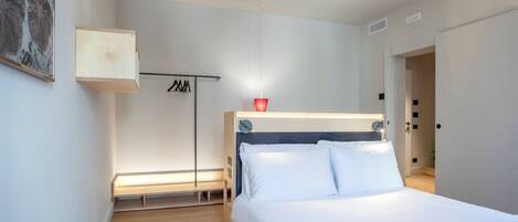 A modern bedroom with a wooden floor, a bed with white linens, a bedside table, a hanging light, and a coat rack.