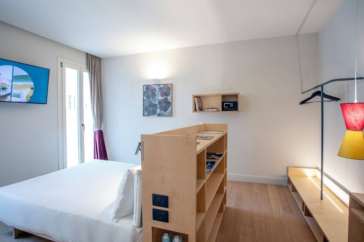 A modern hotel room with a bed, a wooden shelf unit, a TV, and a lamp.