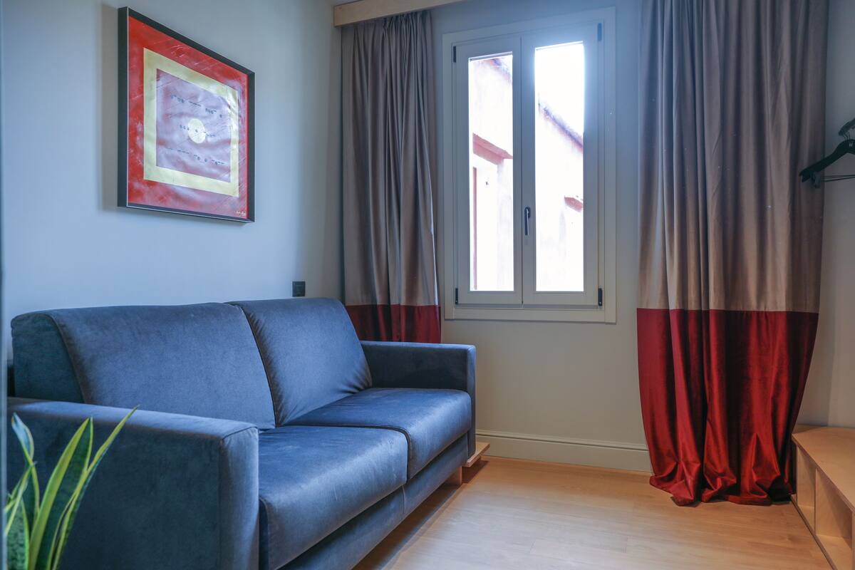 A living room with a blue sofa, a framed picture on the wall, a window with curtains, and a plant.