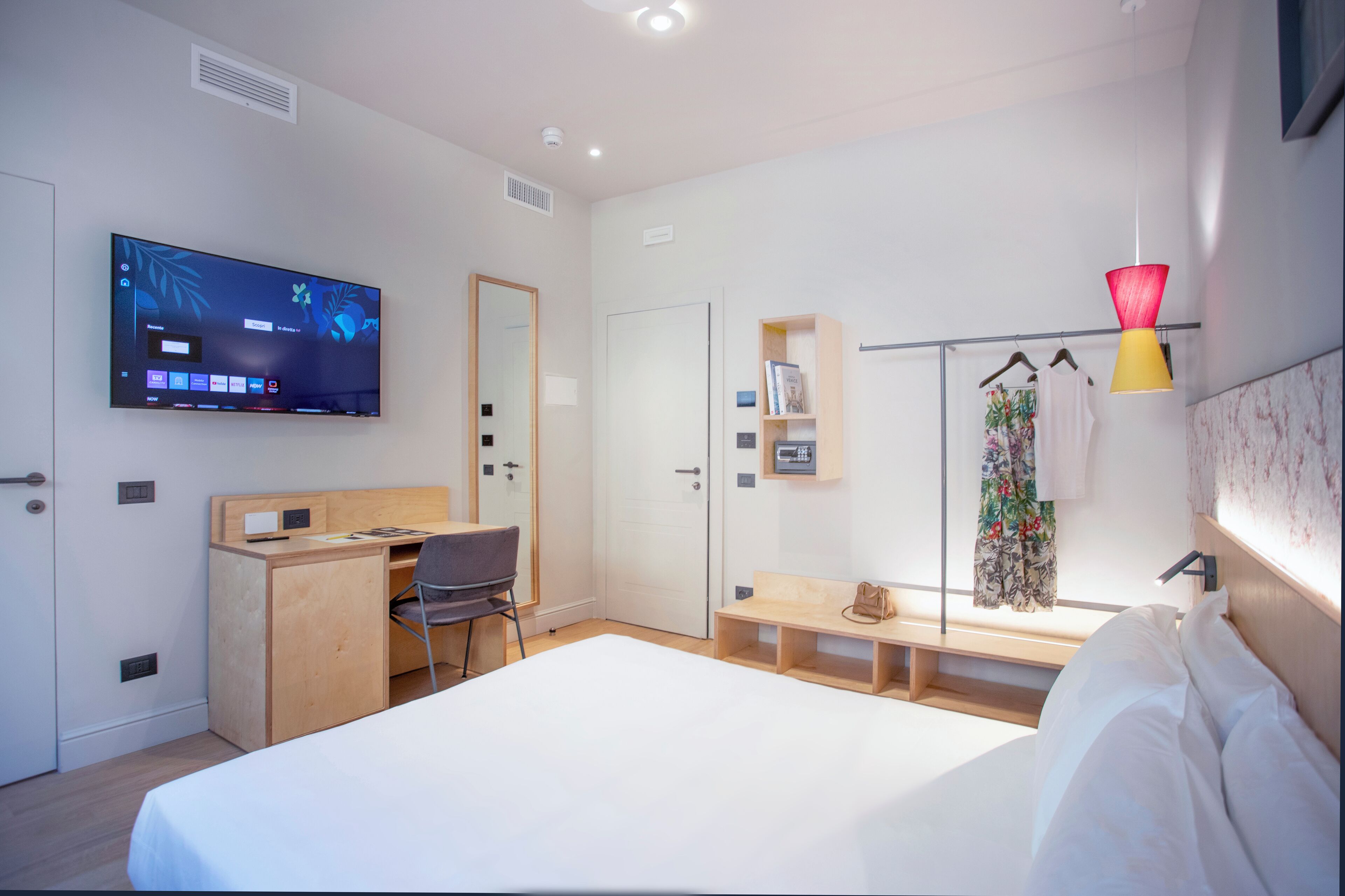 Ein modernes Hotelzimmer mit Bett, Schreibtisch, Stuhl, Fernseher, Kleiderschrank und einer kleinen Ablage mit Büchern und einer Handtasche.