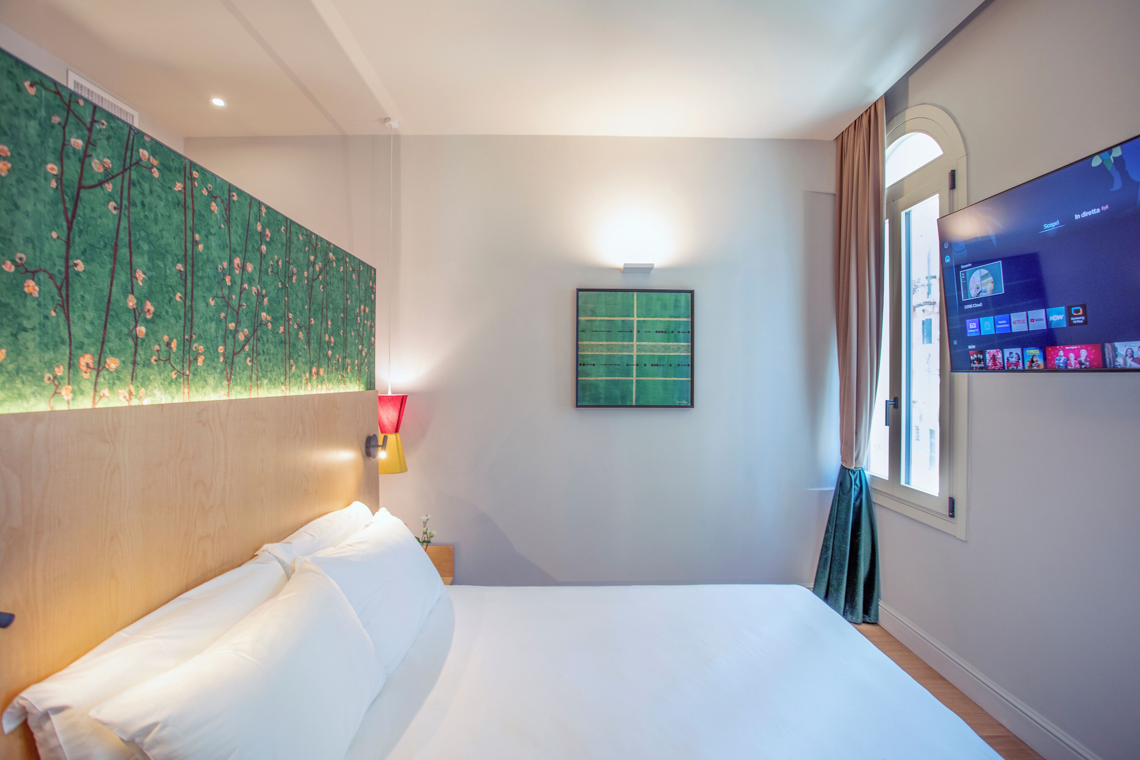 A hotel room with a bed, a painting on the wall, a TV, and a window with curtains.