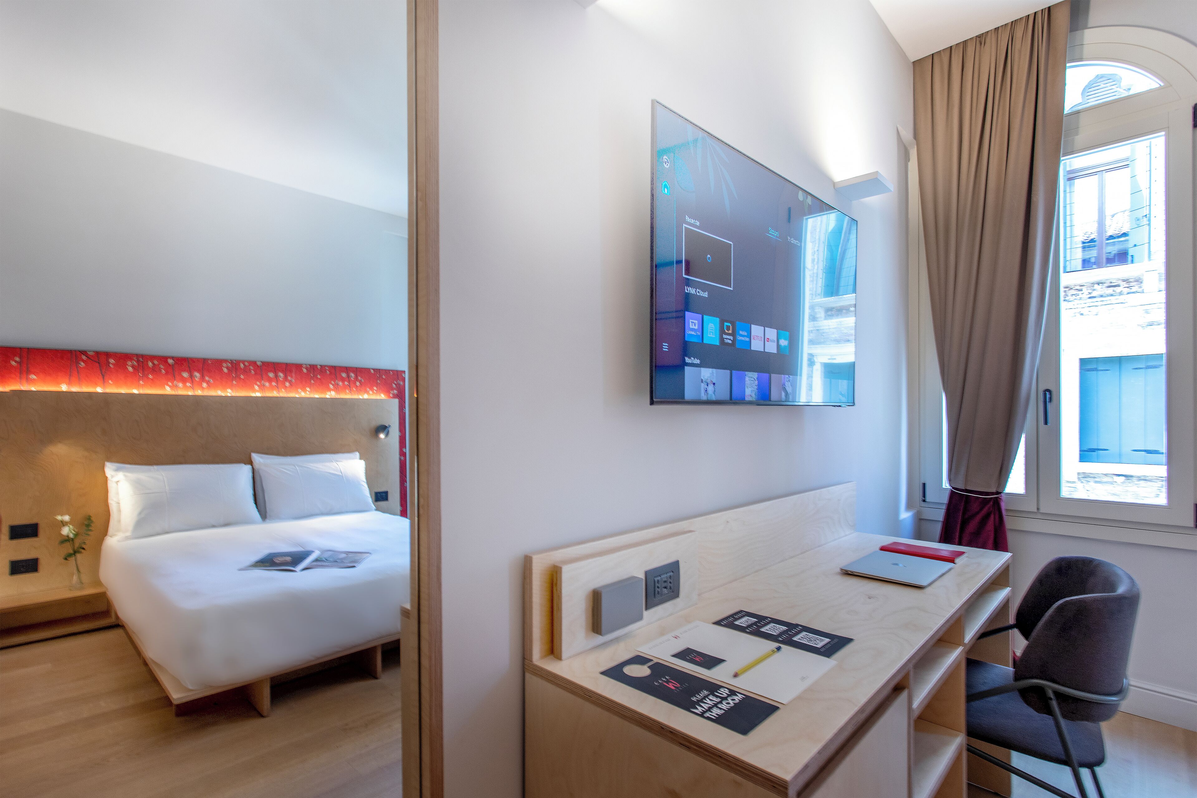 A hotel room with a bed, a desk with a chair, a mounted TV, and a window with curtains.