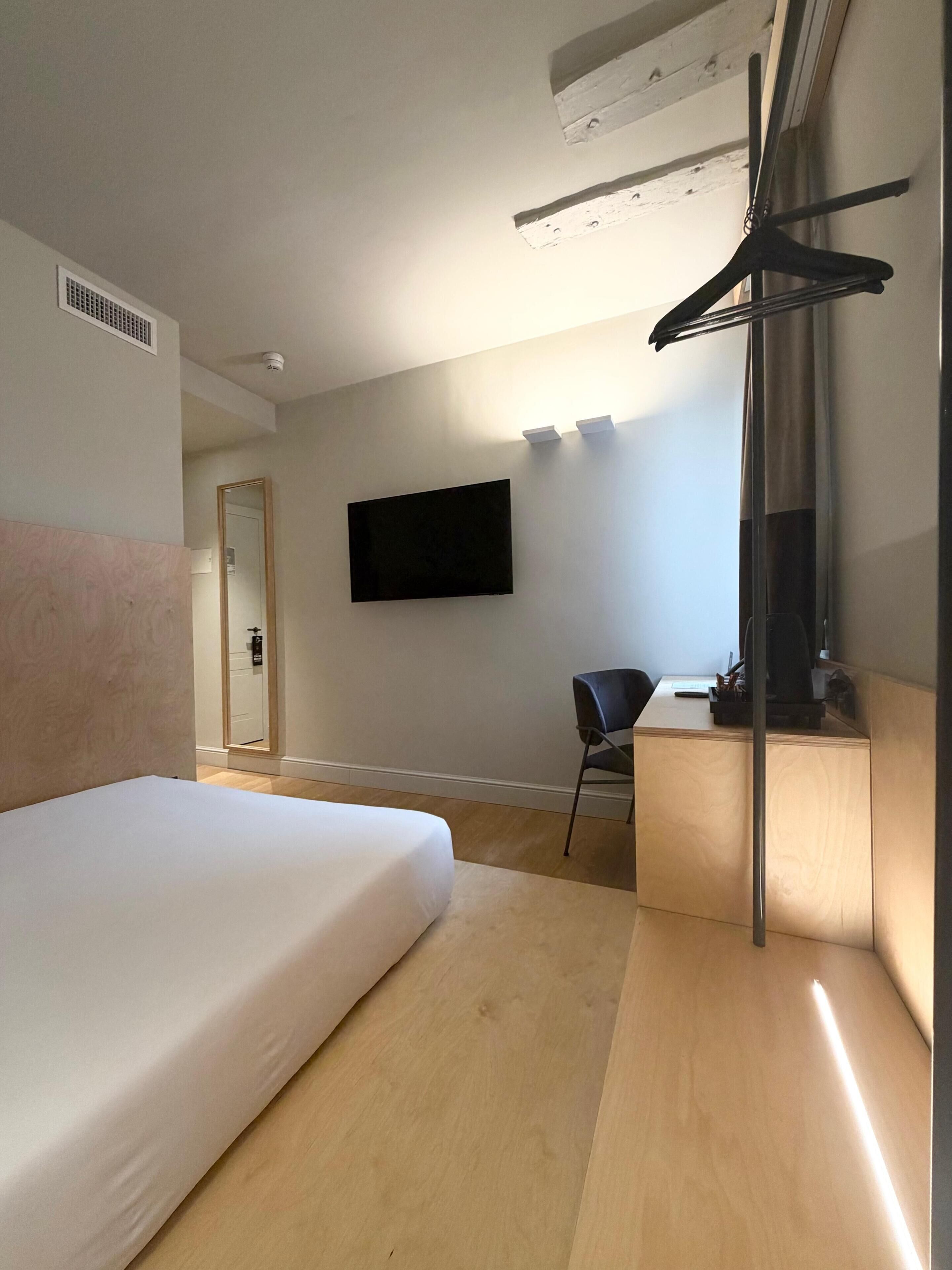 Une chambre d’hôtel équipée d’un lit, d’un bureau, d’une chaise, d’un téléviseur fixé au mur et d’un porte-manteau.