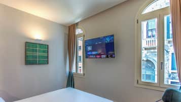 Habitación de hotel con cama, televisor de pantalla plana en la pared, ventana con cortinas y vistas a edificios.