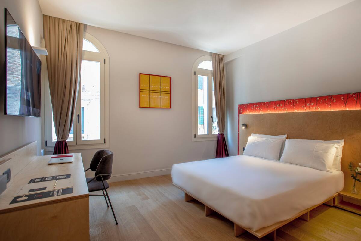 A hotel room with a bed, desk, chair, and two windows with curtains.