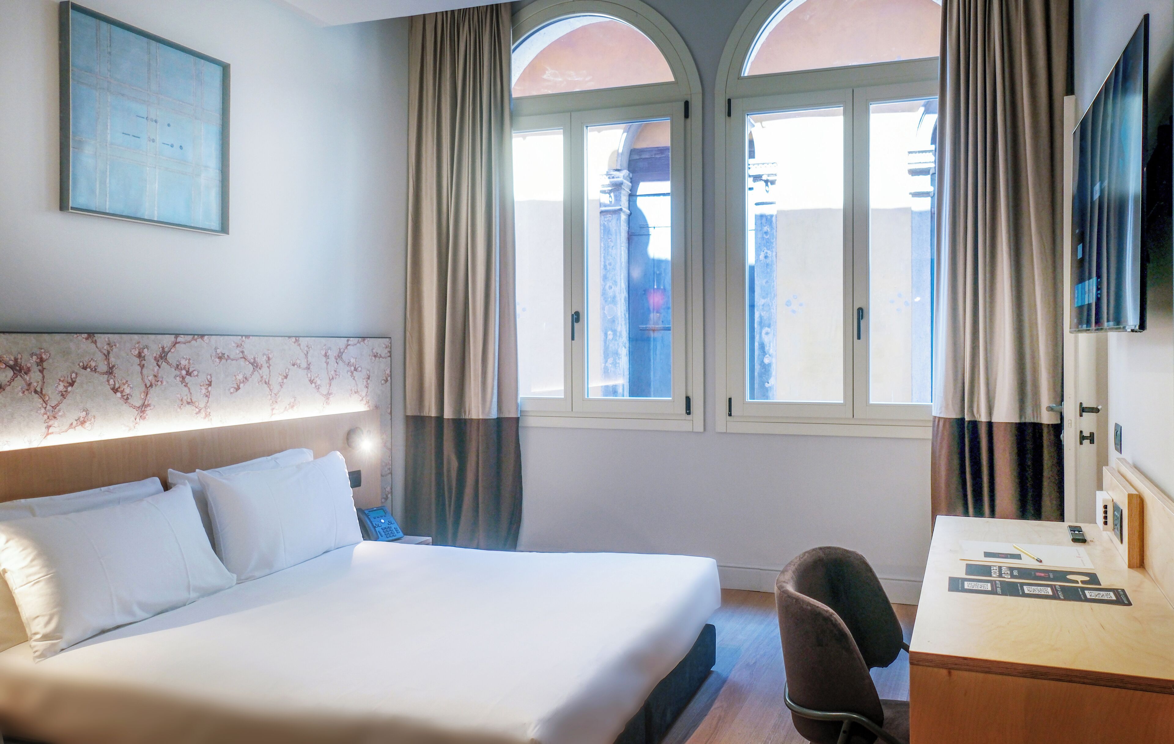 Quarto de hotel com uma cama grande, escrivaninha, cadeira, televisão e janela com cortinas.