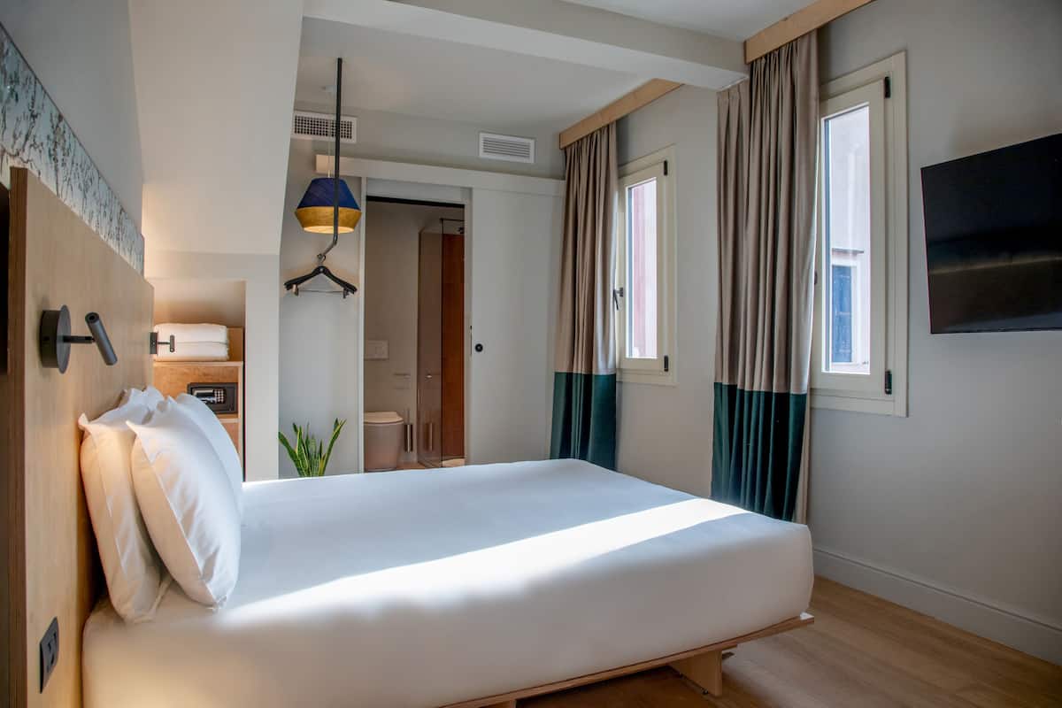 A hotel room with a bed, a wardrobe, a hanging light fixture, a window with curtains, and a television on the wall.