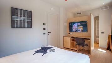A modern hotel room with a bed, a desk with a chair, a television, and a painting on the wall.