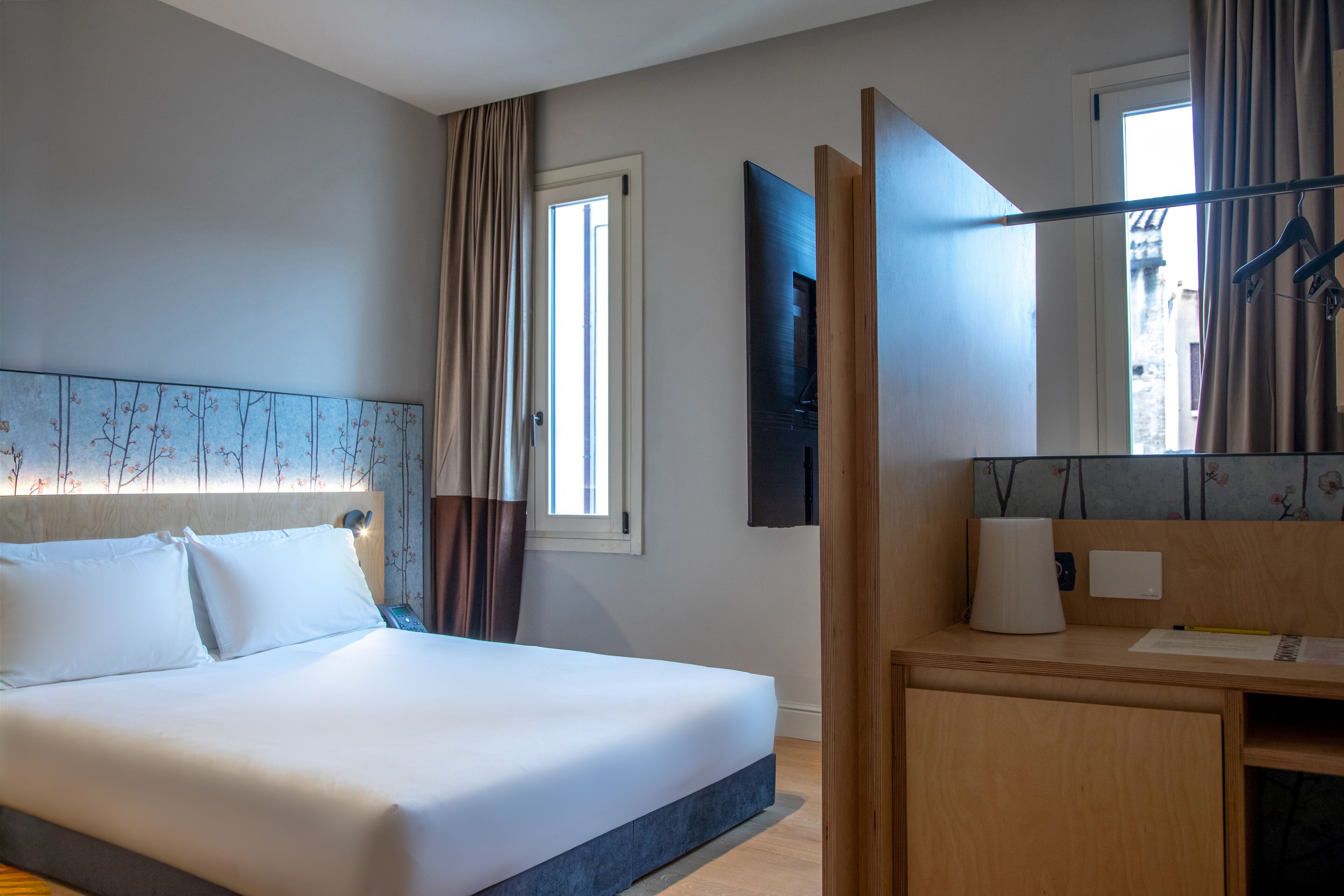 A hotel room with a bed, a desk, a mirror, and a window with curtains.