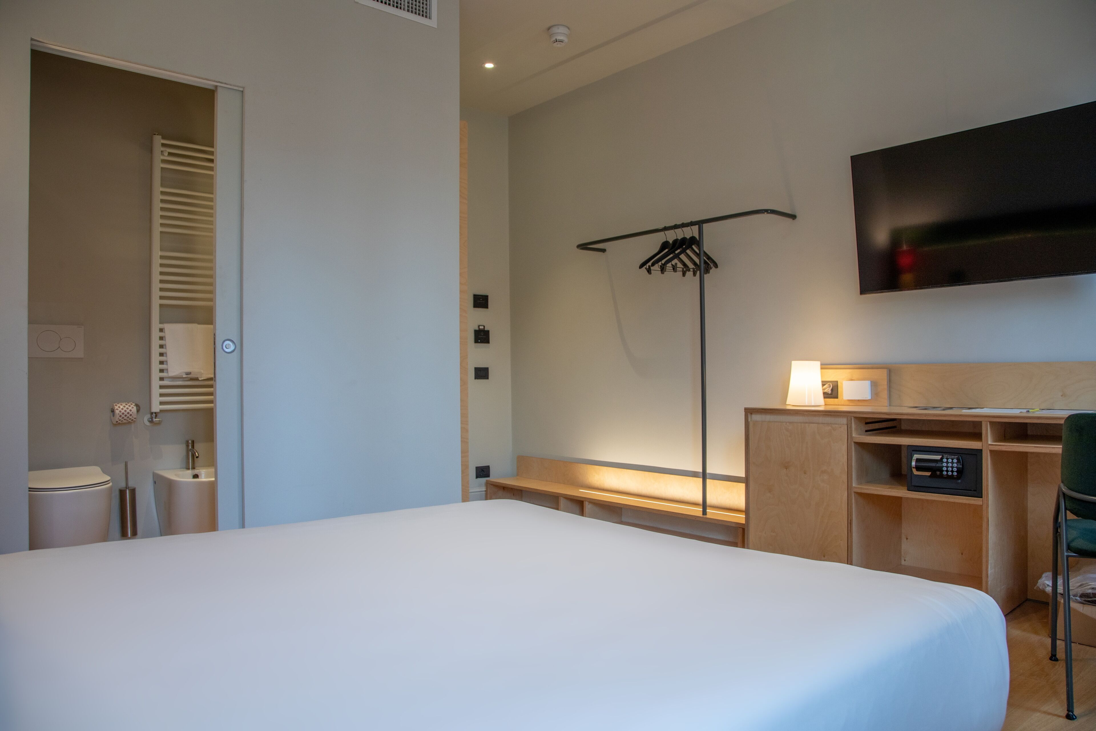 A hotel room with a large bed, a desk with a chair, a TV mounted on the wall, a coat rack, and a small bathroom area.