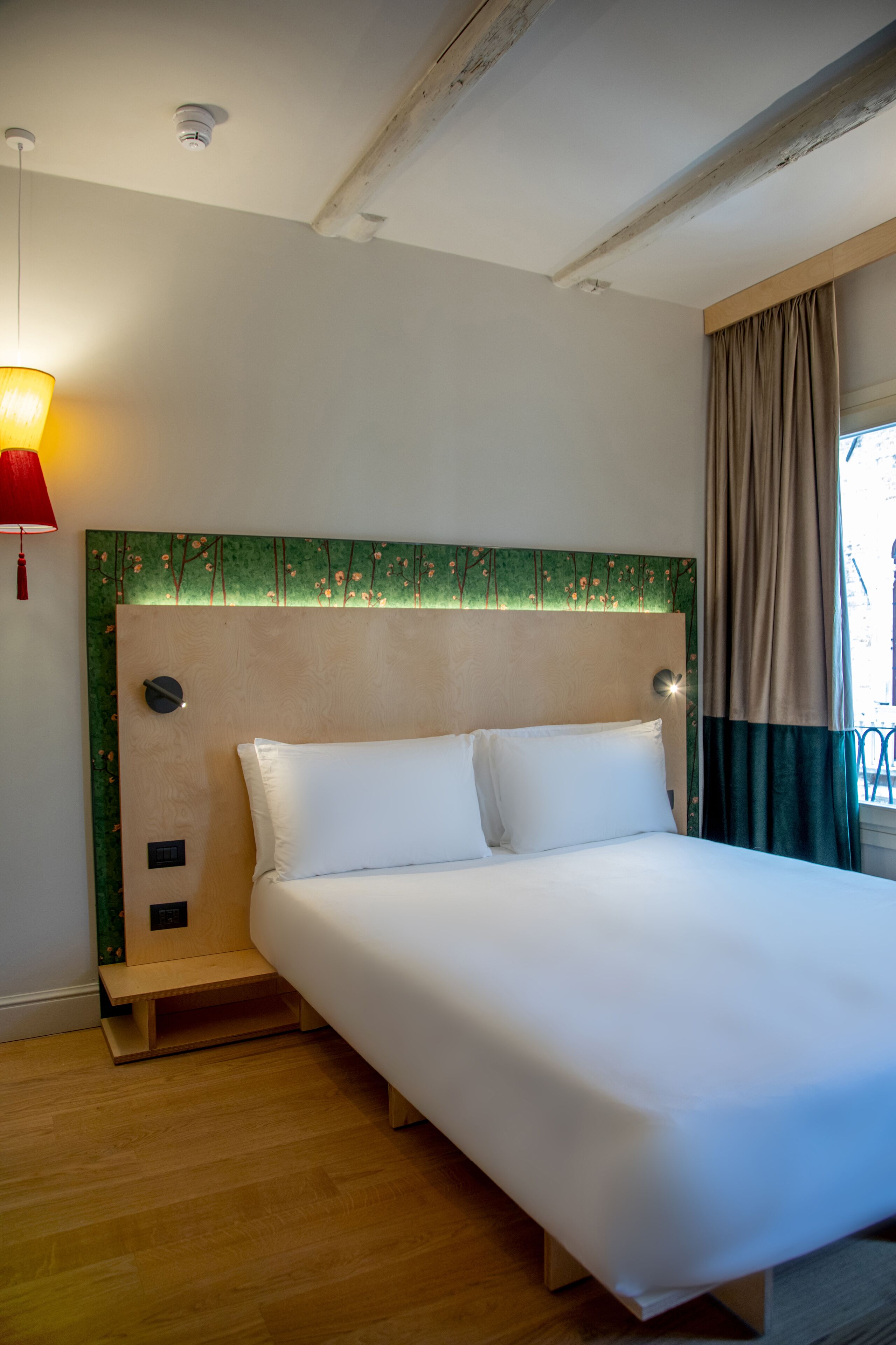 Habitación de hotel con cama, mesita de noche, lámpara y ventana con cortinas.