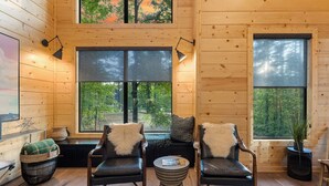A cozy cabin interior with wooden walls, two large windows offering forest views, and a seating area with leather chairs and a small round table.