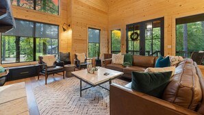 A spacious wooden interior with large windows, a central coffee table, leather sofas, and a fireplace.
