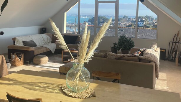 Un loft espaiós amb un sostre alt, zona de menjador amb taula i cadires de fusta, zona d'estar acollidora amb sofà i coixins, i una finestra gran que ofereix vista a la ciutat.