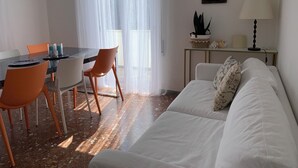 Ein Wohnzimmer mit einem weißen Sofa, einem Esstisch mit orangefarbenen Stühlen und einem Fenster mit transparenten Vorhängen.