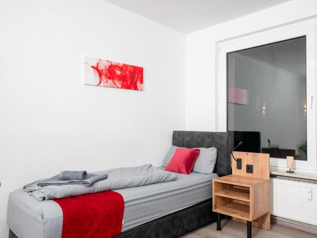 A modern bedroom with a bed, a red and grey blanket, a red pillow, a wooden nightstand, and a large window.