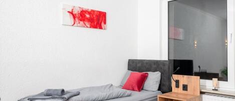 A modern bedroom with a bed, a red and grey blanket, a red pillow, a wooden nightstand, and a large window.