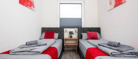 A small, modern room with two beds, each with grey bedding and red pillows. There is a small wooden nightstand between the beds, and a window above it.