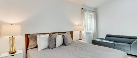 Une chambre à coucher avec un grand lit, deux tables de chevet avec des lampes, un canapé gris et une fenêtre avec des rideaux.