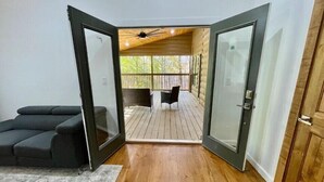 Une pièce avec un plancher en bois, un canapé gris et une vue sur une terrasse extérieure avec une table et des chaises.