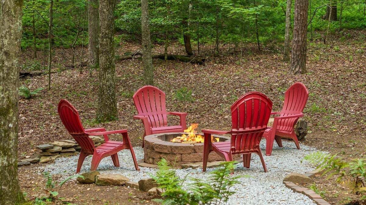 A fire pit with four red Adirondack chairs arranged around it in a forest setting.