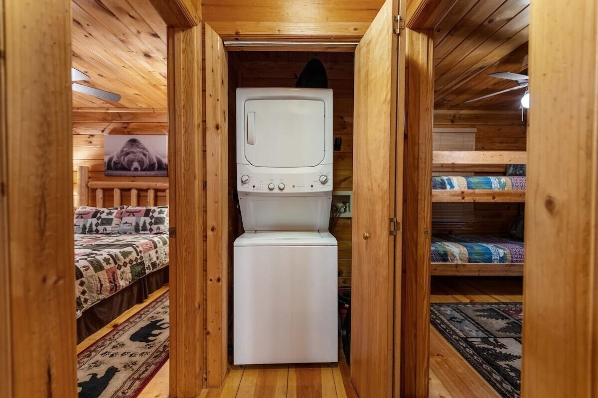 Quarto compacto com decoração em madeira, cama com colcha estampada, eletrodoméstico pequeno branco e televisão sobre suporte.
