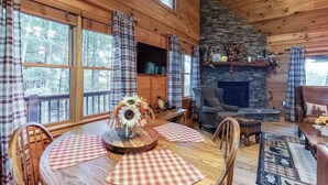 A cozy cabin interior with a stone fireplace, wooden furniture, and large windows overlooking a forest.