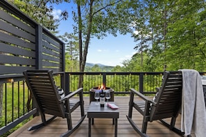 A deck with two chairs, a table, a bottle, and glasses, overlooking a forested area with a fire pit.
