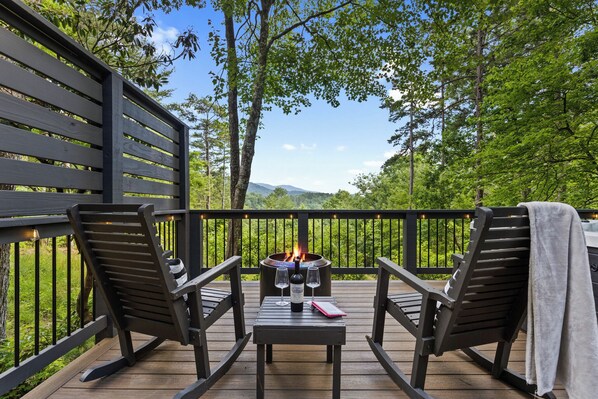 A deck with two chairs, a table, a bottle, and glasses, overlooking a forested area with a fire pit.
