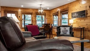 Un acogedor interior de cabaña con un sofá de cuero, una mesa de comedor y una silla con un cojín que dice "ON CABIN TIME".