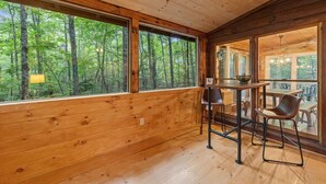 A wooden cabin interior with large windows offering a forest view, a dining area with a table and chairs, and a chandelier. - Wine Knot in Blue Ridge (Blue Ridge)