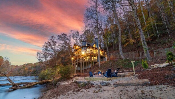 A cabin with a deck overlooking a river, surrounded by trees and a sandy beach.