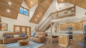 A spacious living area with a high ceiling, wooden beams, a sofa set, a coffee table, and a kitchen with wooden cabinets and appliances.