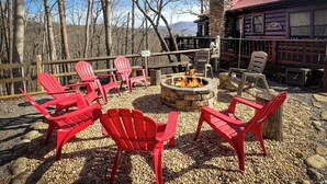 Un espace extérieur rustique aménagé avec des chaises Adirondack rouges disposées autour d’un foyer en pierre, le tout sur fond de forêt et d’une cabane.