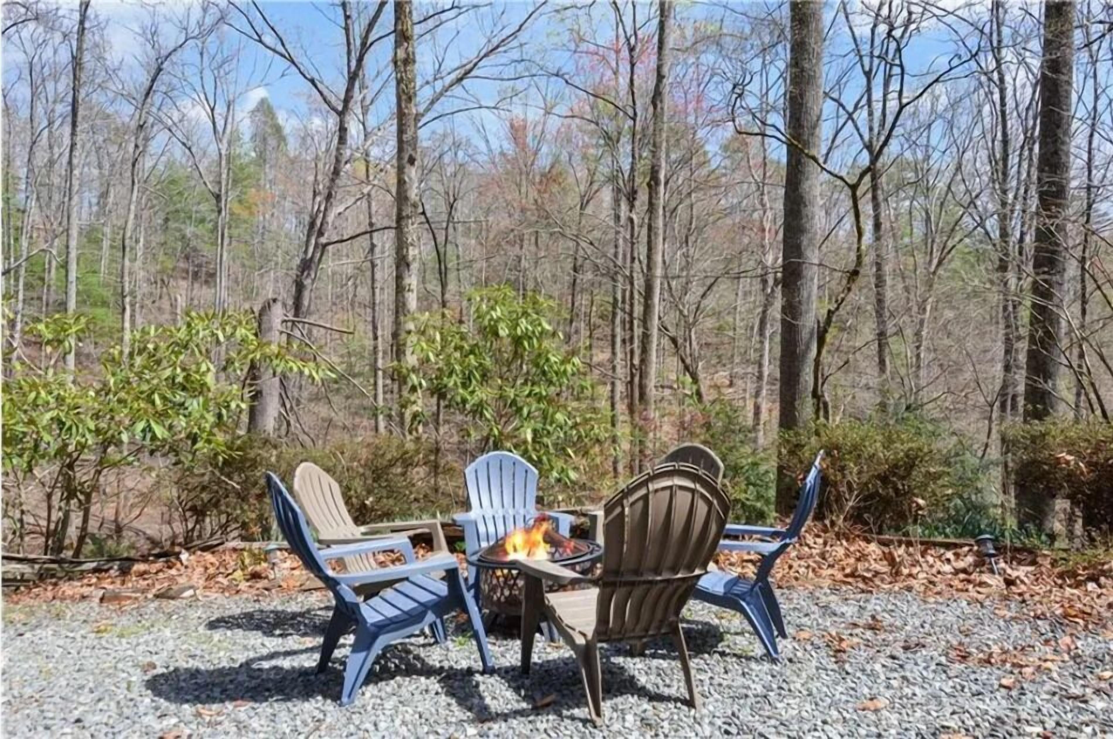 An outdoor seating area with four Adirondack chairs around a small fire pit, surrounded by a forested area.