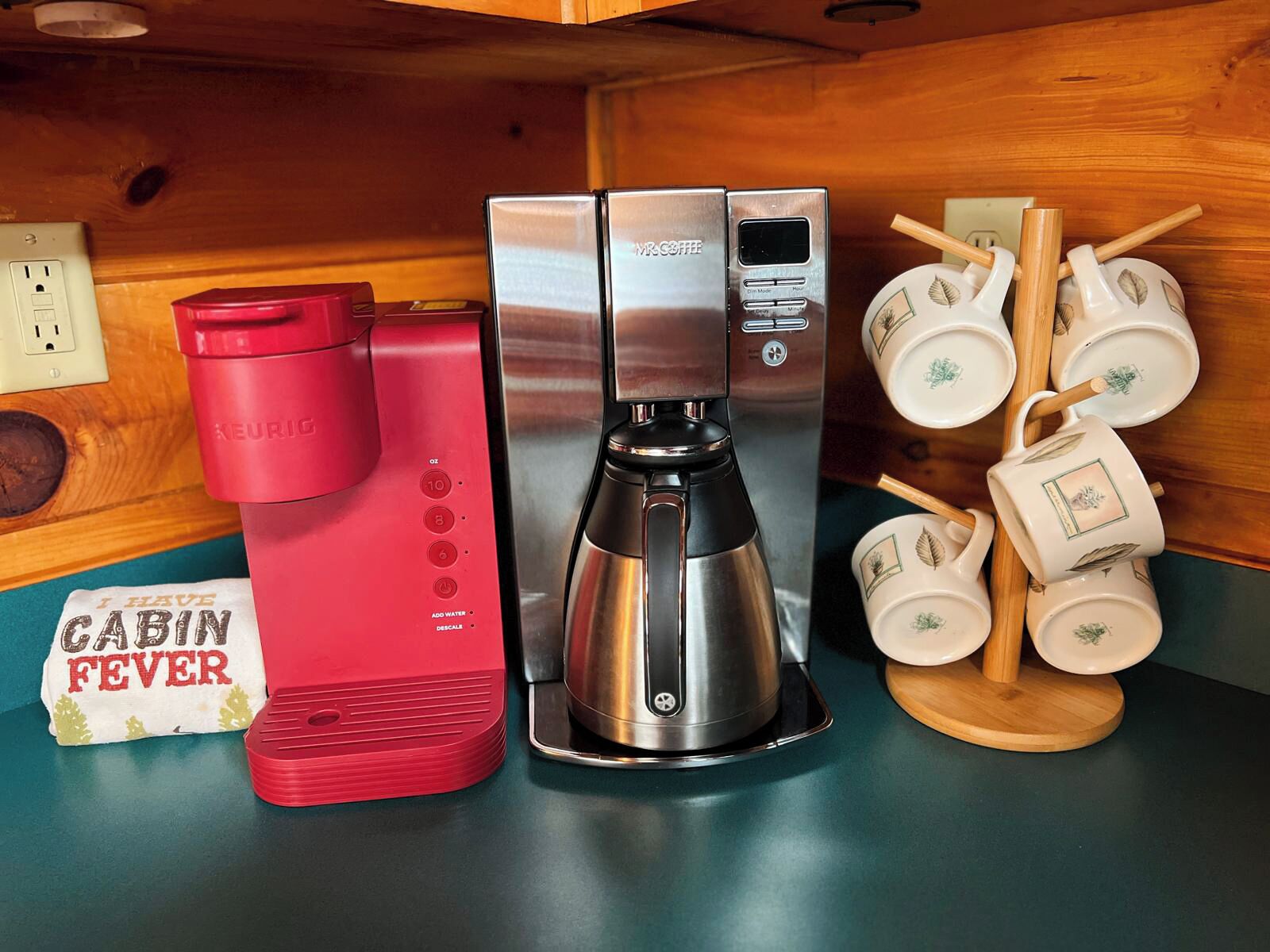 Une machine à café, une bouilloire rouge et un porte-tasses avec des tasses blanches, le tout posé sur un comptoir en bois.