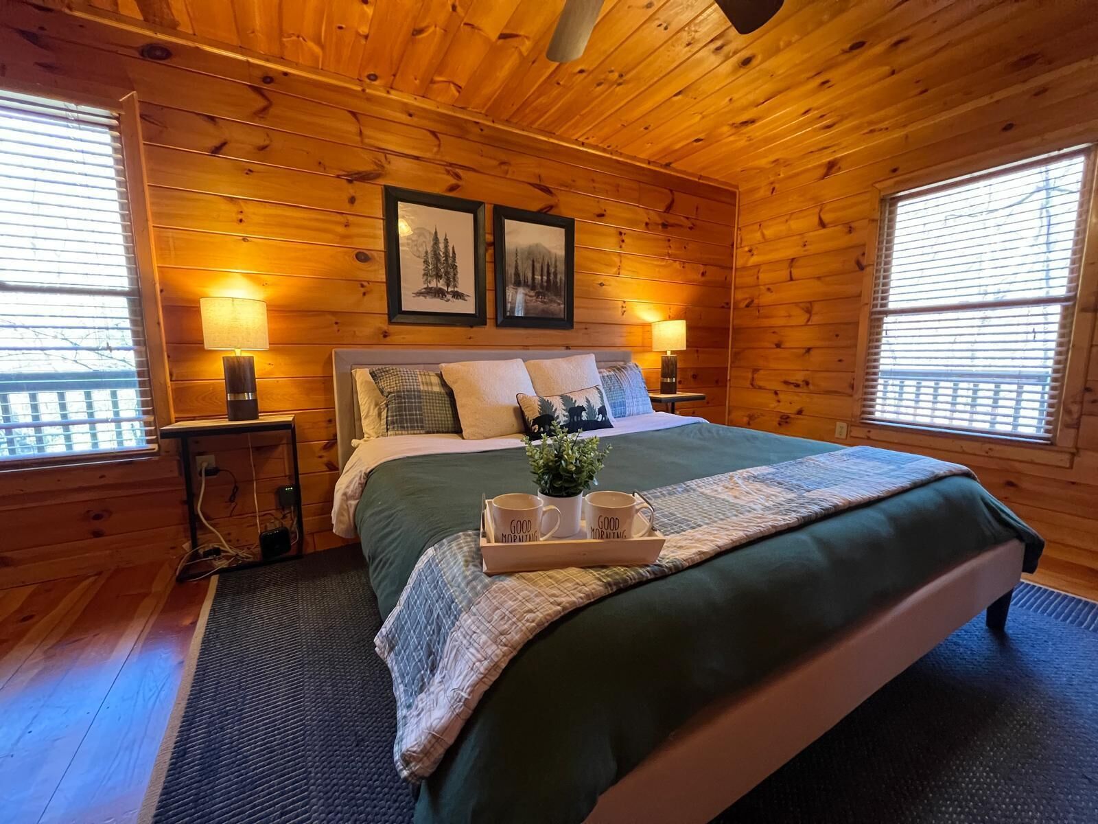 A wooden cabin bedroom with a bed, two bedside lamps, framed pictures on the wall, and a tray with mugs on the bed.