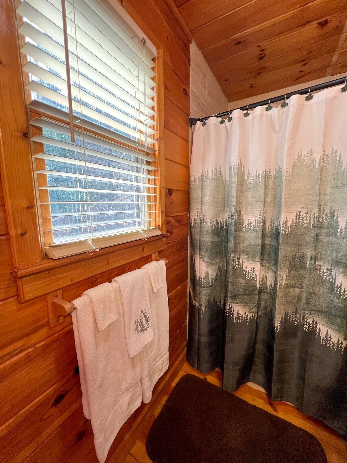 A wooden cabin interior with a window, a curtain with a forest pattern, and a towel on a hook.